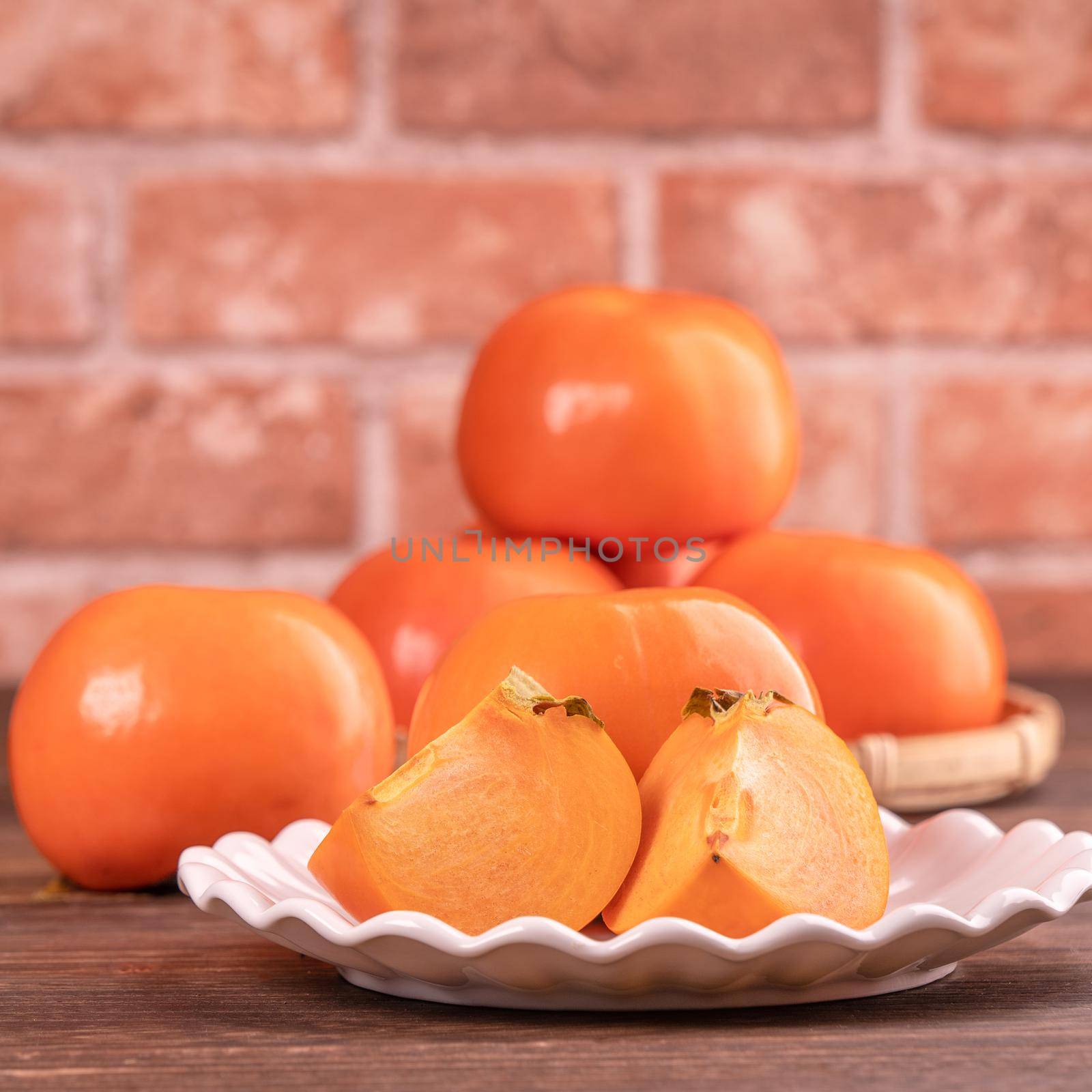 Sliced sweet persimmon kaki in a bamboo sieve basket on dark wooden table with red brick wall background, Chinese lunar new year fruit design concept, close up.