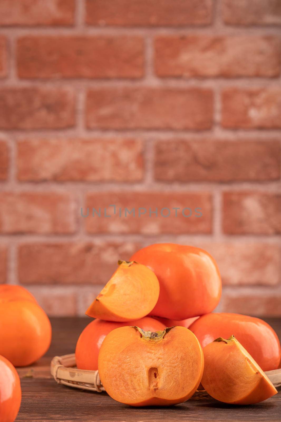 Sliced sweet persimmon kaki in a bamboo sieve basket on dark wooden table with red brick wall background, Chinese lunar new year fruit design concept, close up.
