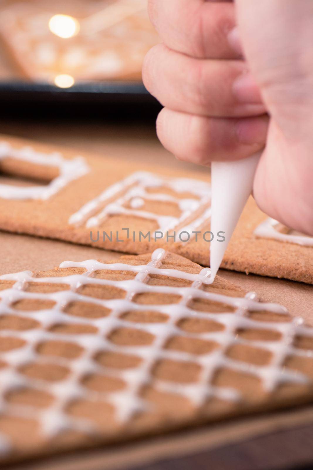 Woman is decorating gingerbread cookies house with white frosting icing cream topping on wooden table background, baking paper in kitchen, close up, macro. by ROMIXIMAGE