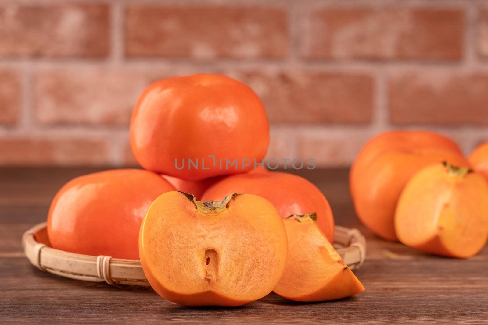 Sliced sweet persimmon kaki in a bamboo sieve basket on dark wooden table with red brick wall background, Chinese lunar new year fruit design concept, close up.
