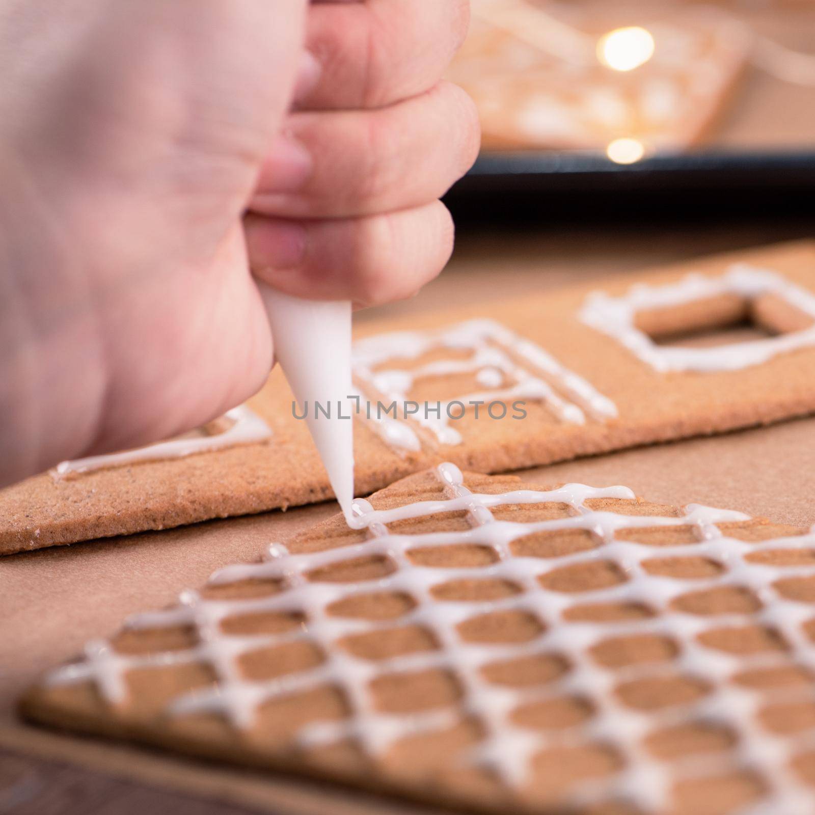 Woman is decorating gingerbread cookies house with white frosting icing cream topping on wooden table background, baking paper in kitchen, close up, macro.