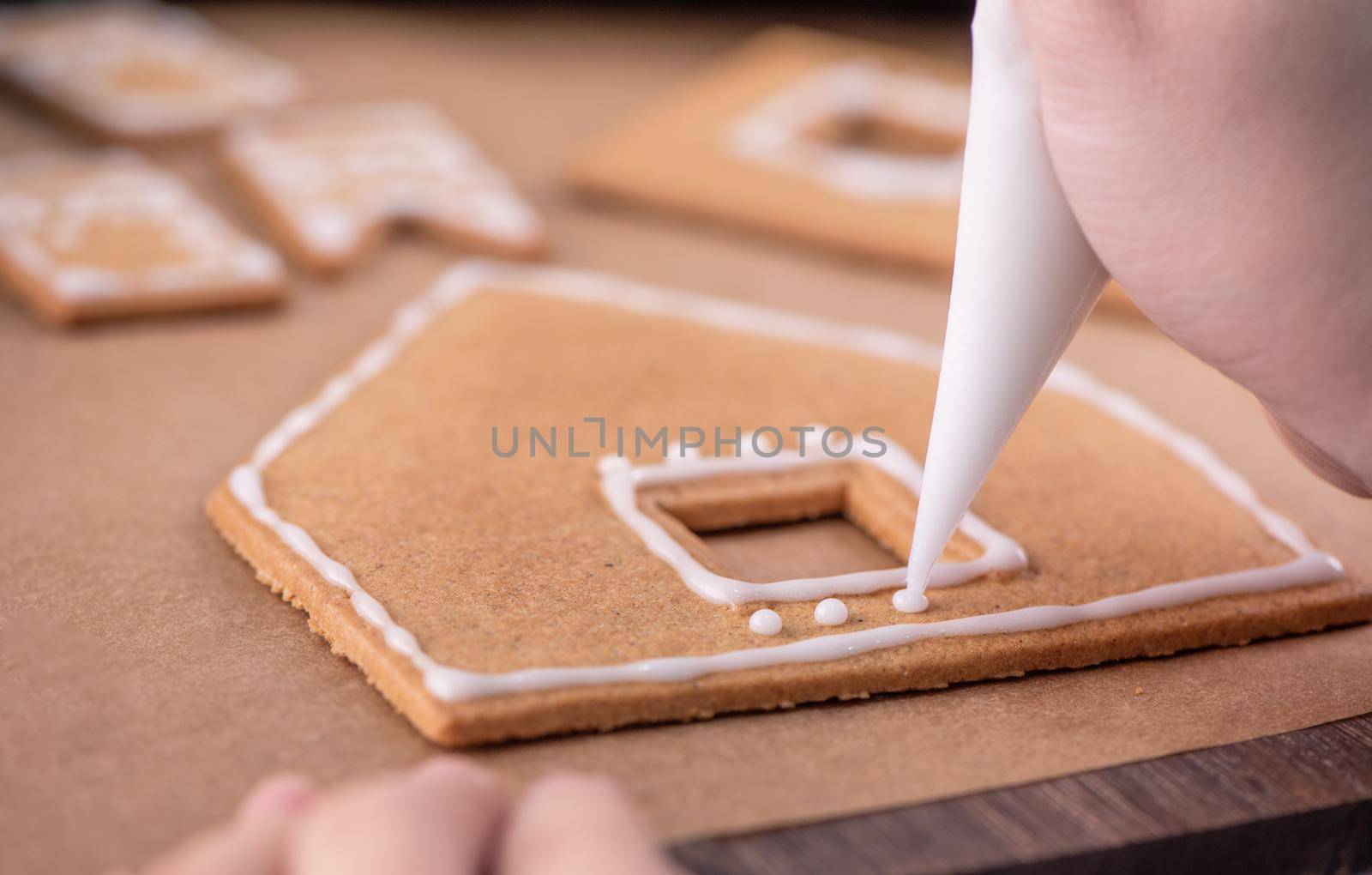 Woman is decorating gingerbread cookies house with white frosting icing cream topping on wooden table background, baking paper in kitchen, close up, macro.