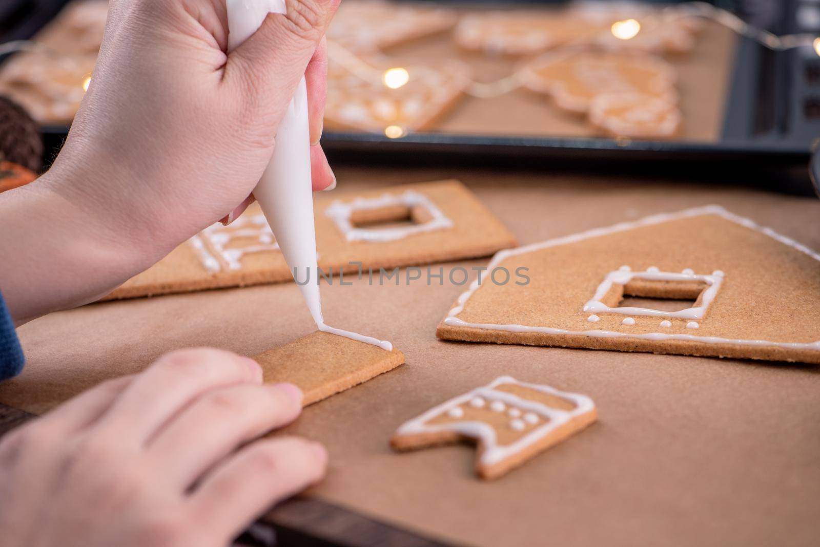 Woman is decorating gingerbread cookies house with white frosting icing cream topping on wooden table background, baking paper in kitchen, close up, macro.