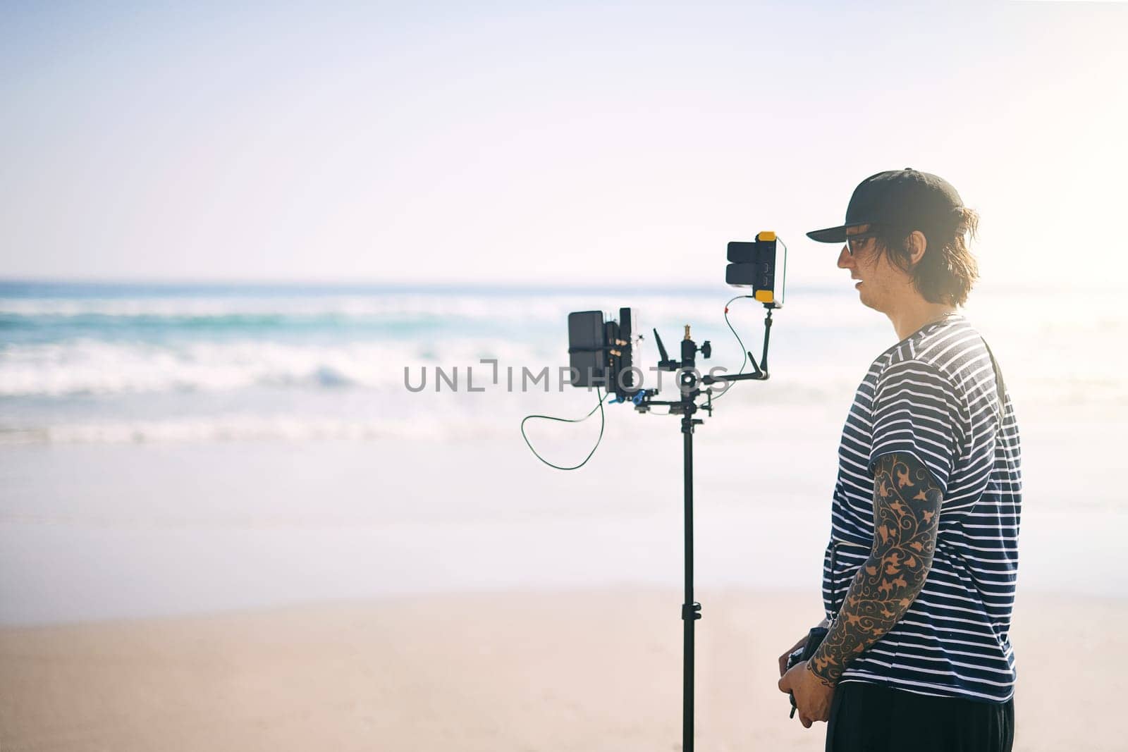 This going to be a long day. a focused young man shooting a scene with a state of the art video camera outside on a beach during the day. by YuriArcurs