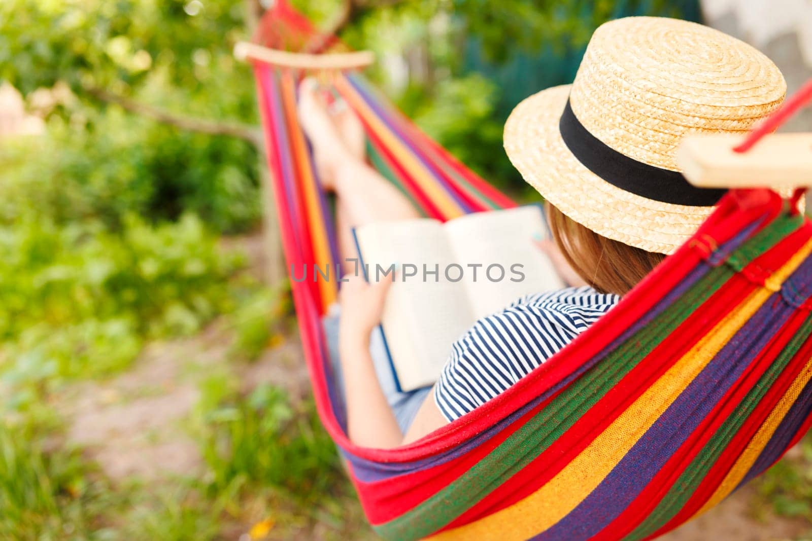 Young woman reading book while lying in comfortable hammock at green garden.