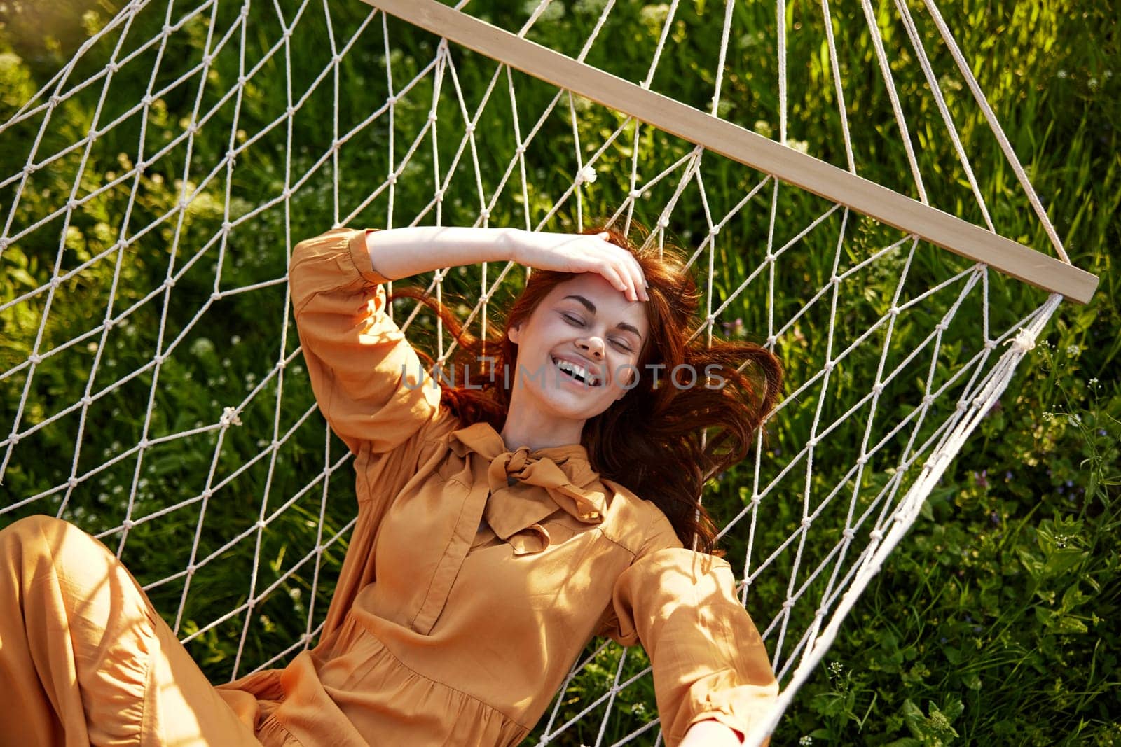 a beautiful, elegant woman lies in a long orange dress on a mesh hammock resting in nature, illuminated by the warm sunset light, holding her hand on her head, covering her face from the sun. High quality photo