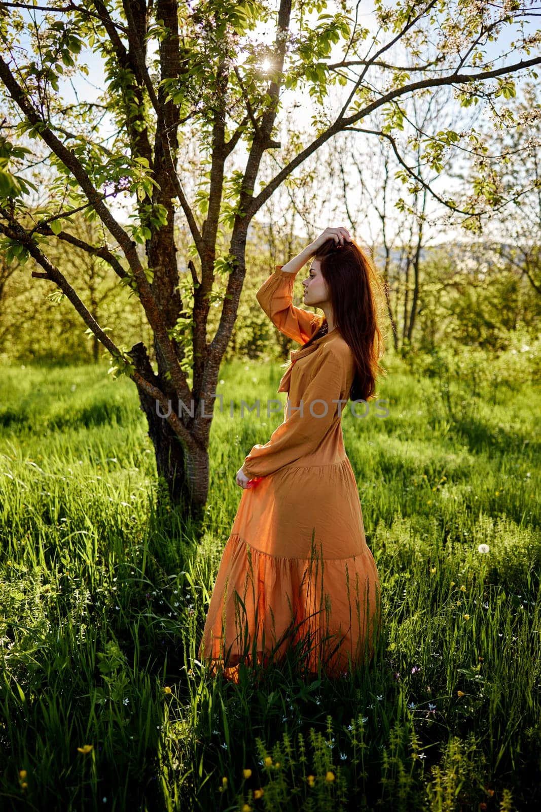 a lovely woman stands in nature in a bright, long orange dress, illuminated from the back by the sunset rays of the sun and holds her skirt with one hand, and straightens her hair with the other by Vichizh