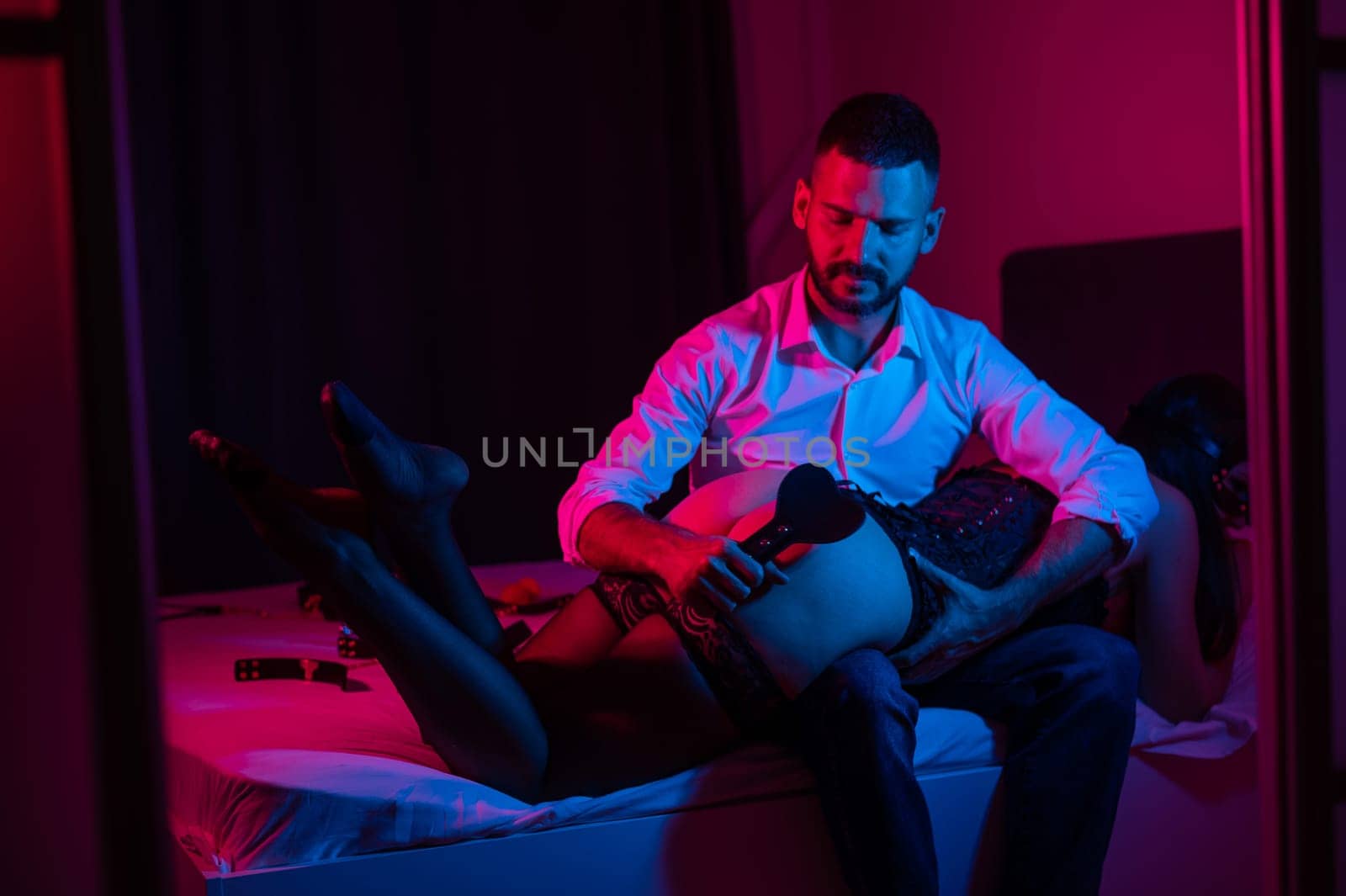 Bearded caucasian man spanks a woman with a leather whip while sitting on a bed in neon light. BDSM sex preferences