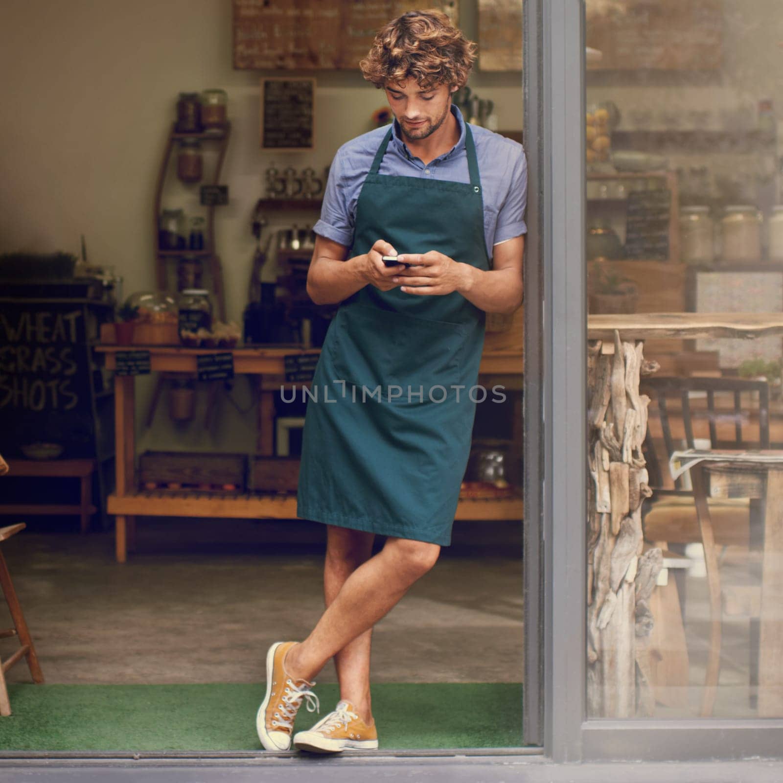 Coffee shop, typing and phone of a man as a small business owner at front door. Entrepreneur person as barista, manager or waiter in restaurant with mobile app for service, marketing or communication by YuriArcurs