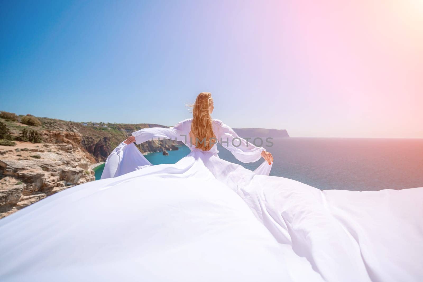 woman sea white dress. Blonde with long hair on a sunny seashore in a white flowing dress, rear view, silk fabric waving in the wind. Against the backdrop of the blue sky and mountains on the seashore. by Matiunina