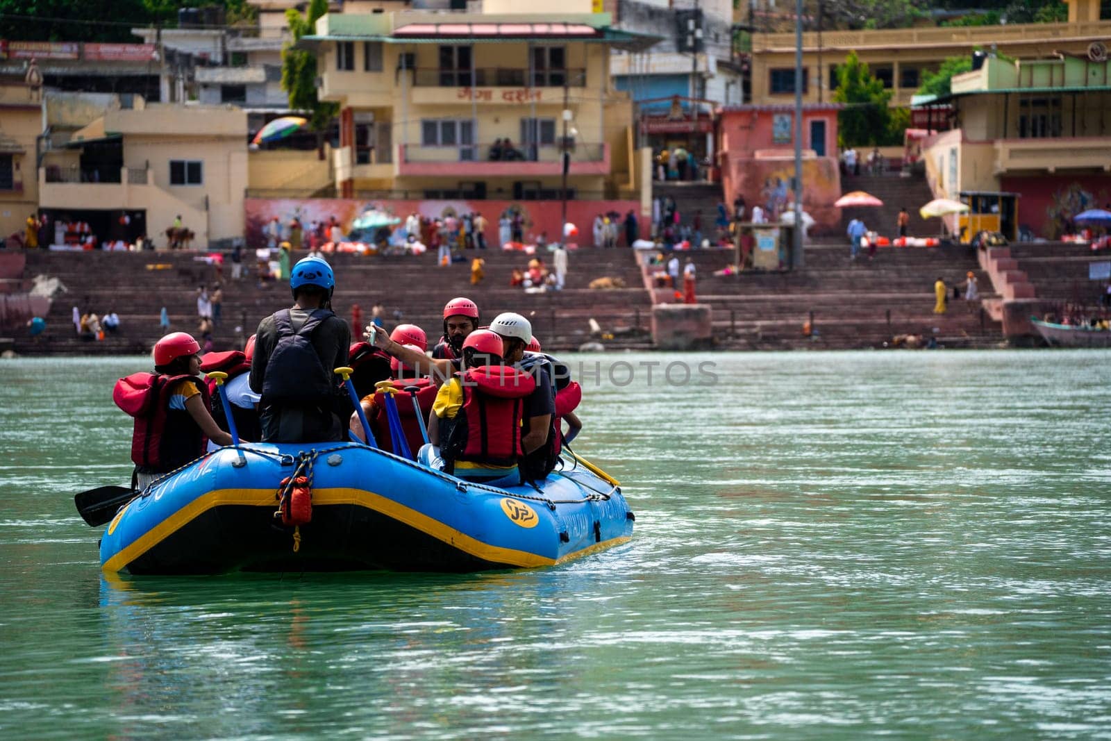 Rishikesh, Haridwar, India - circa 2023: family friends sitting in raft enjoying adventure sports while crossing in front of ghat and temple on the banks of the holy river ganga