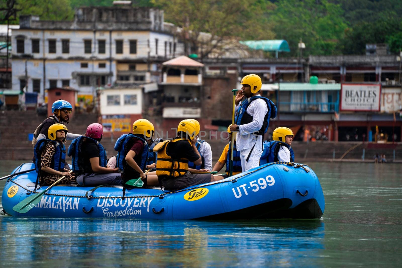 Rishikesh, Haridwar, India - circa 2023: man standing in the front of inflatable white water raft showing leadership with team in helmets and life jackets on river ganga in front of janki setu bridge with himalaya in distance