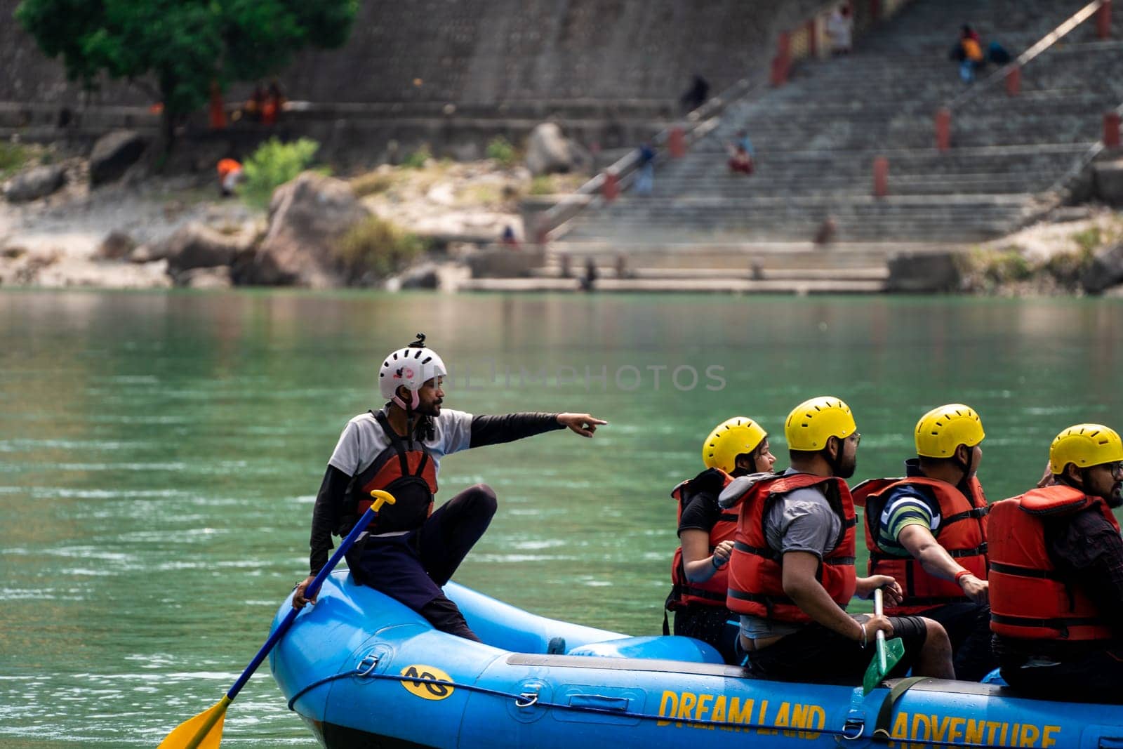 Rishikesh, Haridwar, India - circa 2023: man standing in the front of inflatable white water raft showing leadership with team in helmets and life jackets on river ganga in front of janki setu bridge with himalaya in distance