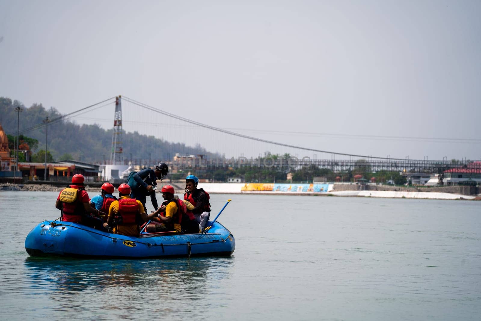 Rishikesh, Haridwar, India - circa 2023: family friends in white water raft in front of temples ghats and the ram setu bridge in holy tourist town of rishikesh