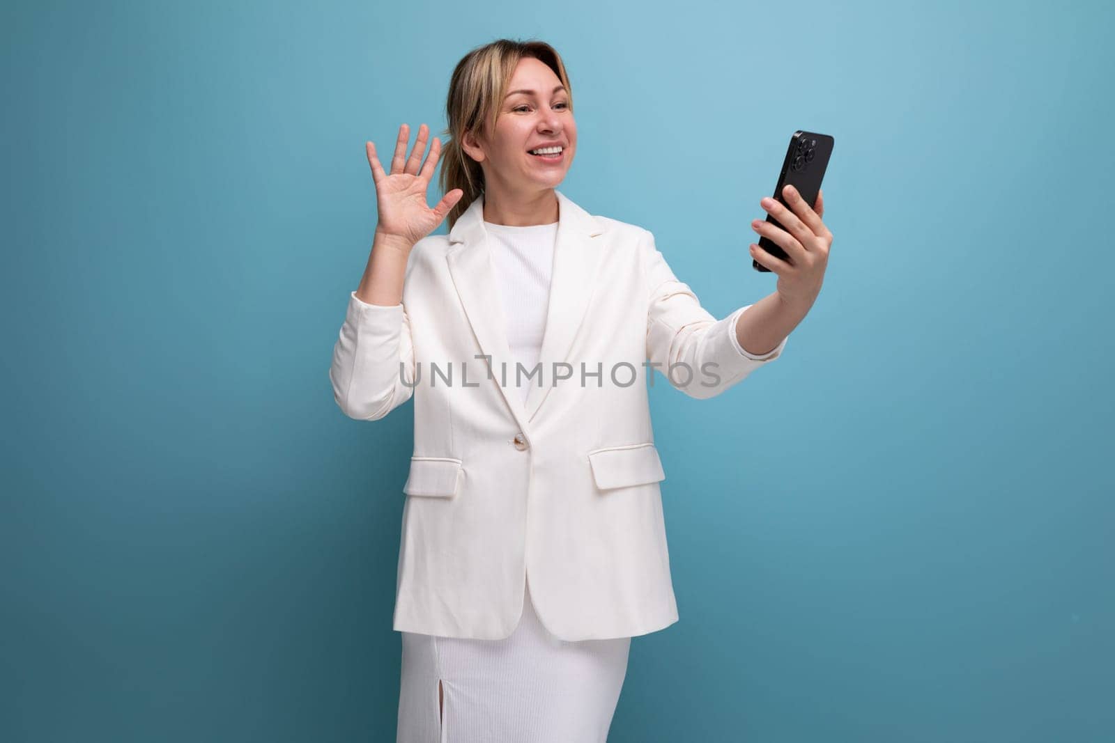 young well-groomed slender blond woman wearing a white jacket and dress uses a smartphone.