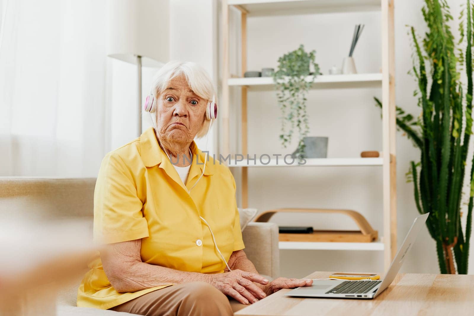 An elderly woman wearing headphones with a laptop sitting on the couch at home and working in a yellow shirt in front of a window, surprise, the lifestyle of a pensioner. High quality photo