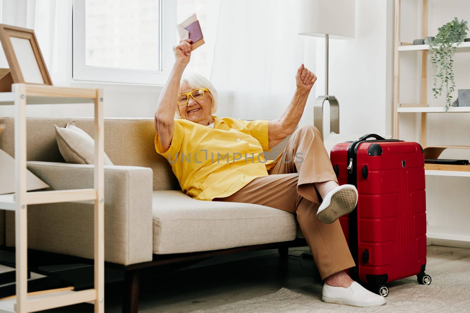 Happy senior woman with passport and travel ticket packed a red suitcase, vacation and health care. Smiling old woman joyfully sitting on the sofa before the trip raised her hands up in joy. High quality photo