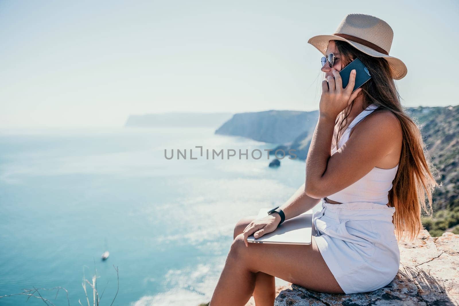 Successful business woman in yellow hat working on laptop by the sea. Pretty lady typing on computer at summer day outdoors. Freelance, travel and holidays concept.