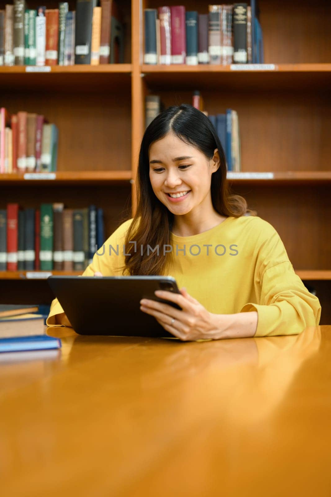 Smiling female university student using digital tablet, doing research, learning lessons, preparing for exams in library.