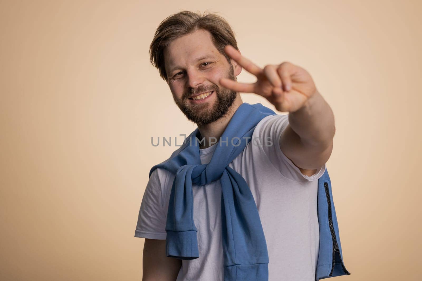 Young caucasian man showing victory sign, hoping for success and win, doing peace gesture, smiling with kind optimistic expression. Handsome bearded guy isolated alone on beige studio background