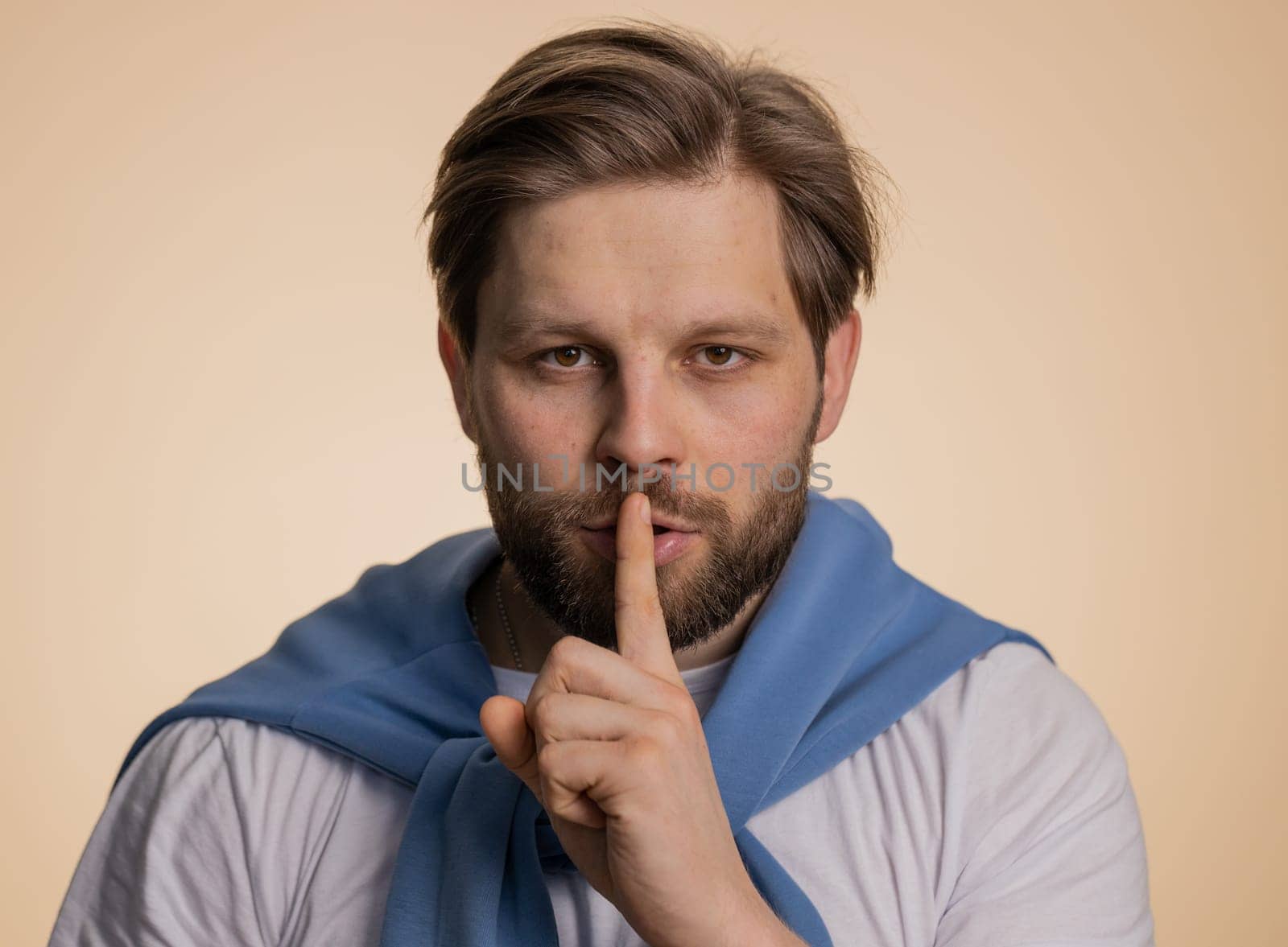 Shh be quiet please. Young caucasian man presses index finger to lips makes silence gesture sign do not tells secret, stop gossip. Handsome bearded guy isolated alone on beige studio wall background