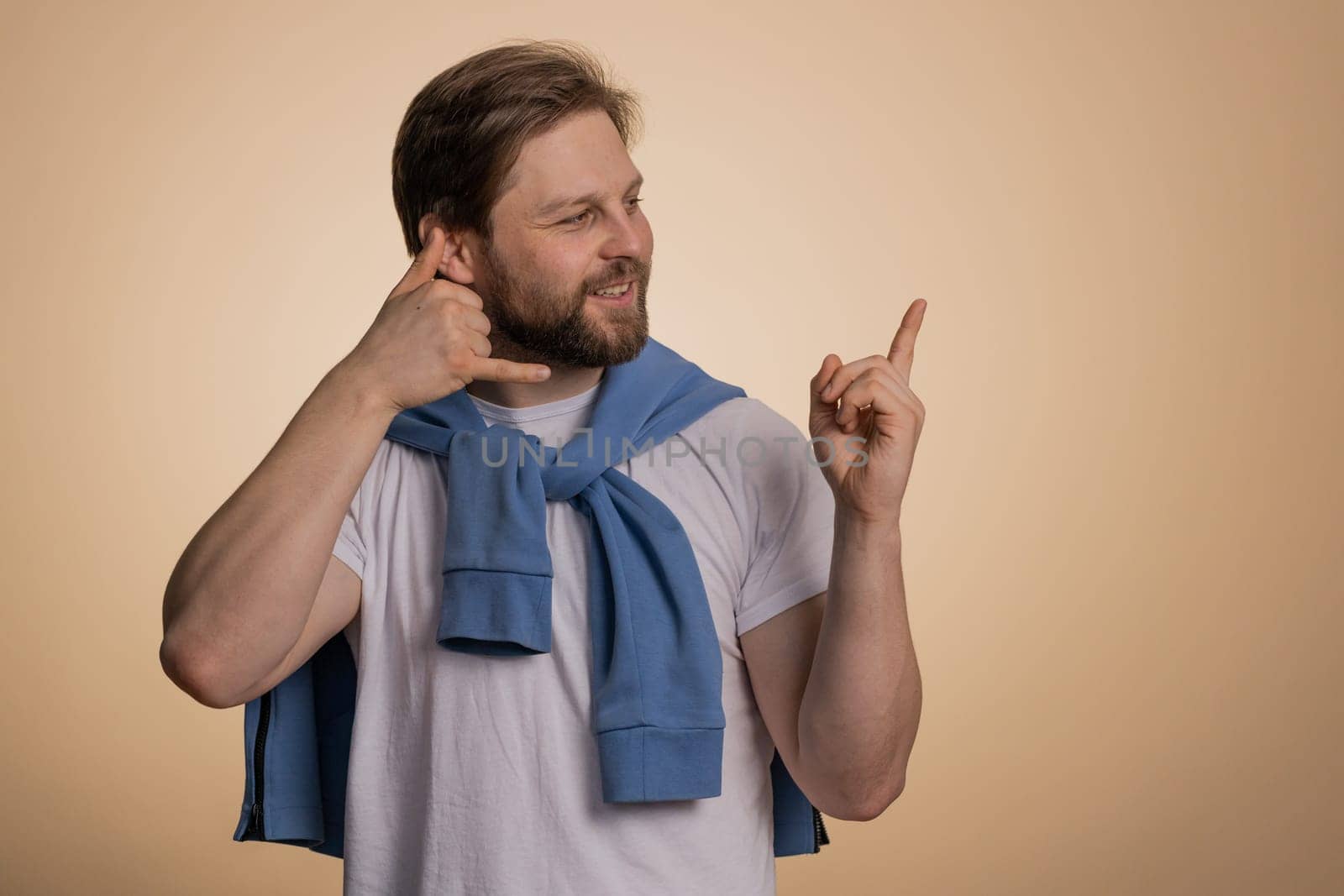 Call us, here is contact number. Handsome bearded caucasian man in t-shirt looking at camera doing phone gesture like says hey you call me back conversation. Guy isolated on beige background, indoor