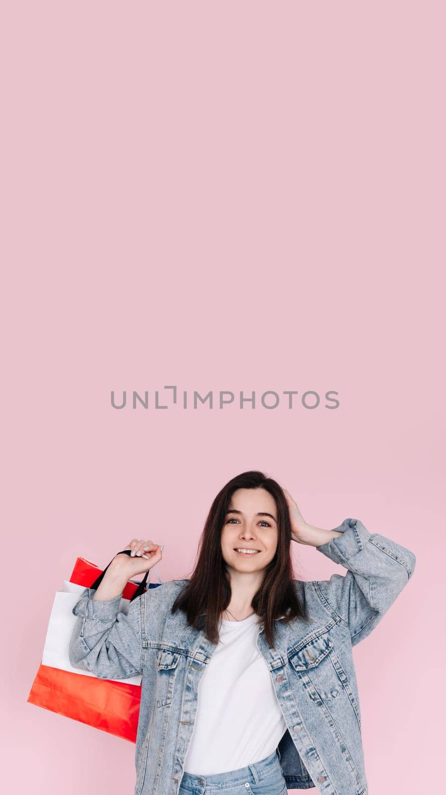 Excited young woman in casual denim shirt raises her arm in celebration, standing in front of a pink background, happy after shopping or finding a great deal online. Vertical photography