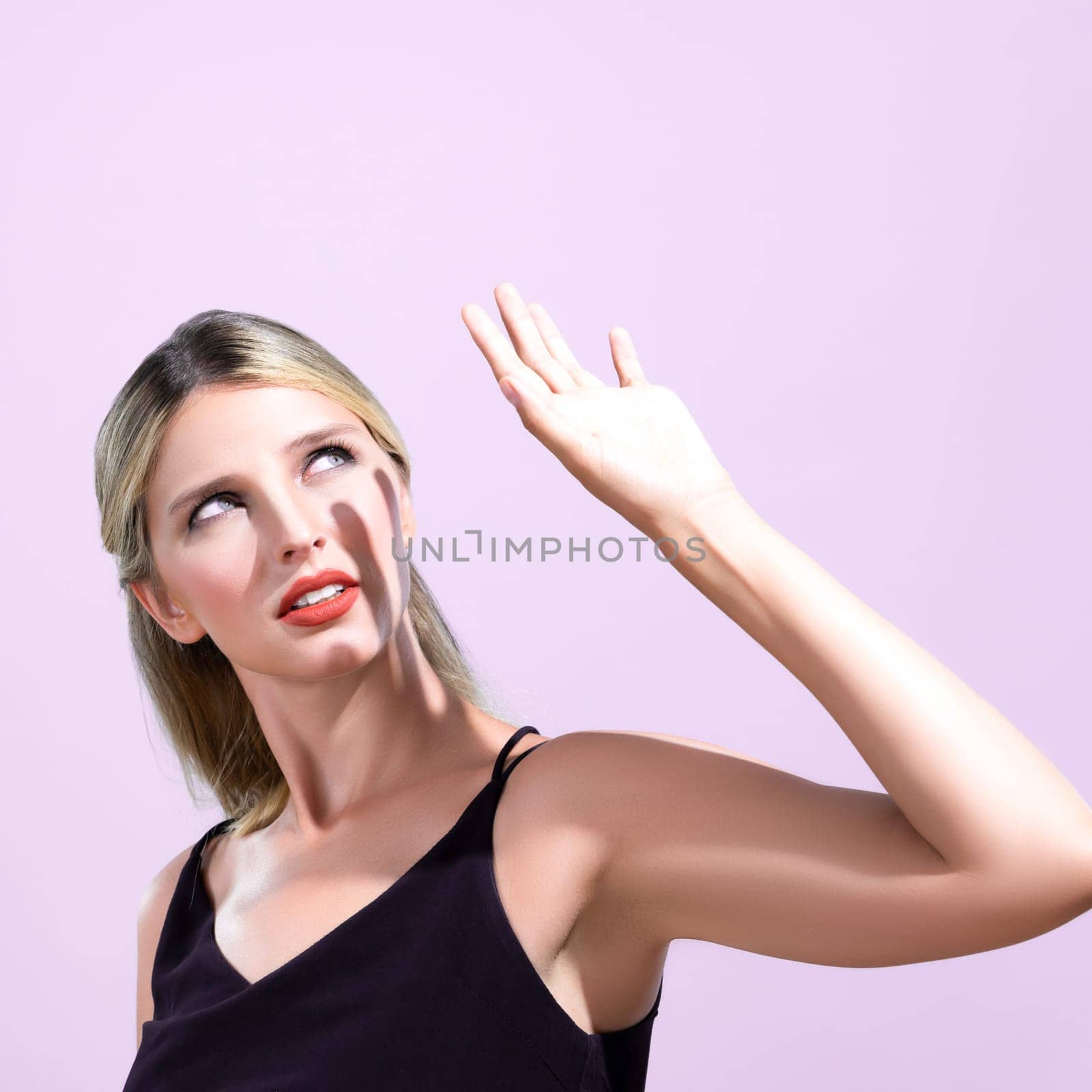 Closeup young blond hair woman with perfect skin and soft alluring facial makeup raise her hand cover her face from bright sunlight in pink isolated background for skincare sunscreen product.