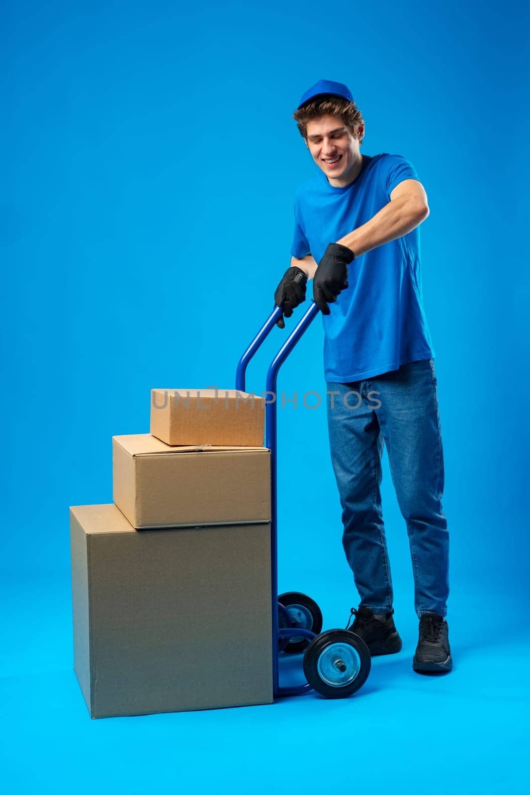 Young happy delivery man carrying boxes on truck against blue background in studio