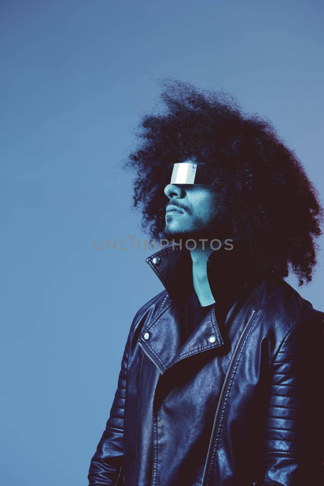 Portrait of fashion man with curly hair with stylish glasses on blue background multinational, colored light, black leather jacket trend, modern concept. High quality photo