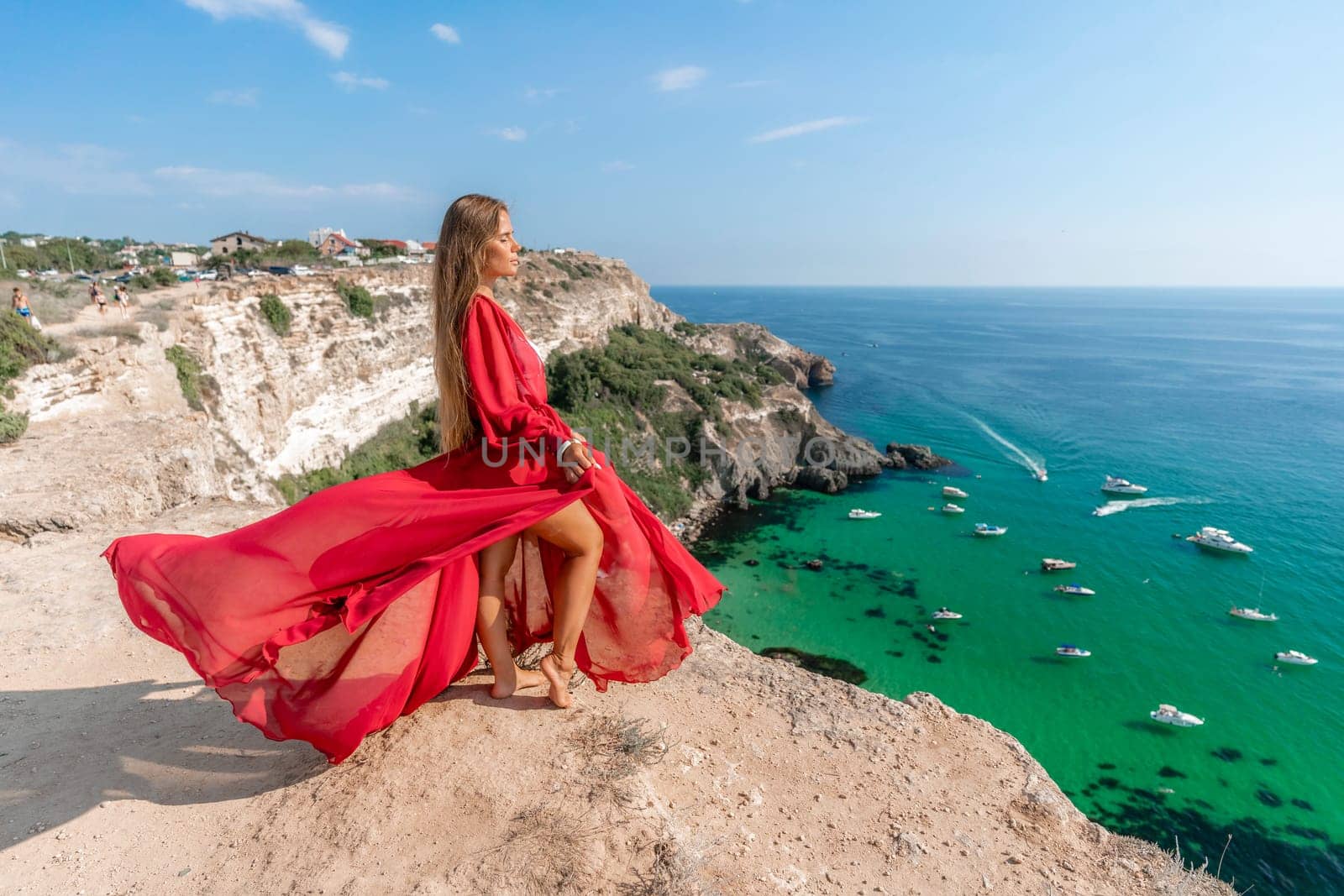 Red Dress Woman sea Cliff. A beautiful woman in a red dress and white swimsuit poses on a cliff overlooking the sea on a sunny day. Boats and yachts dot the background. by Matiunina