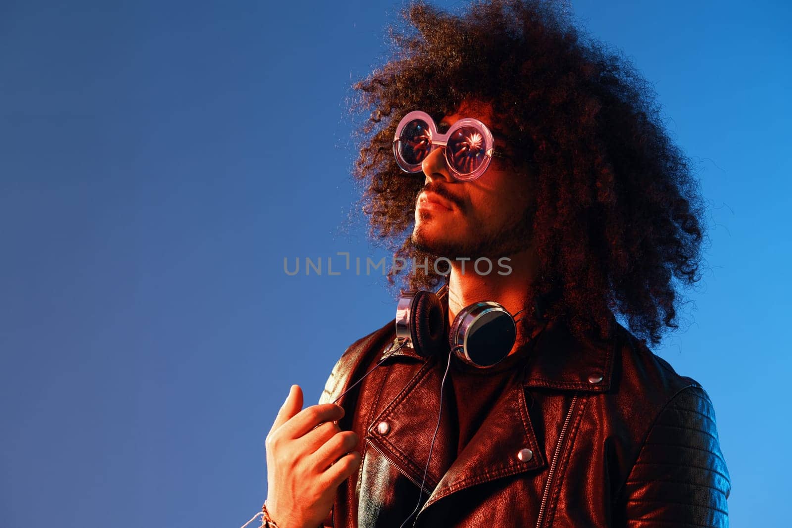 Portrait of a stylish man with curly hair with glasses and headphones on a blue background multinational, colored light, black leather jacket trend, modern concept. by SHOTPRIME