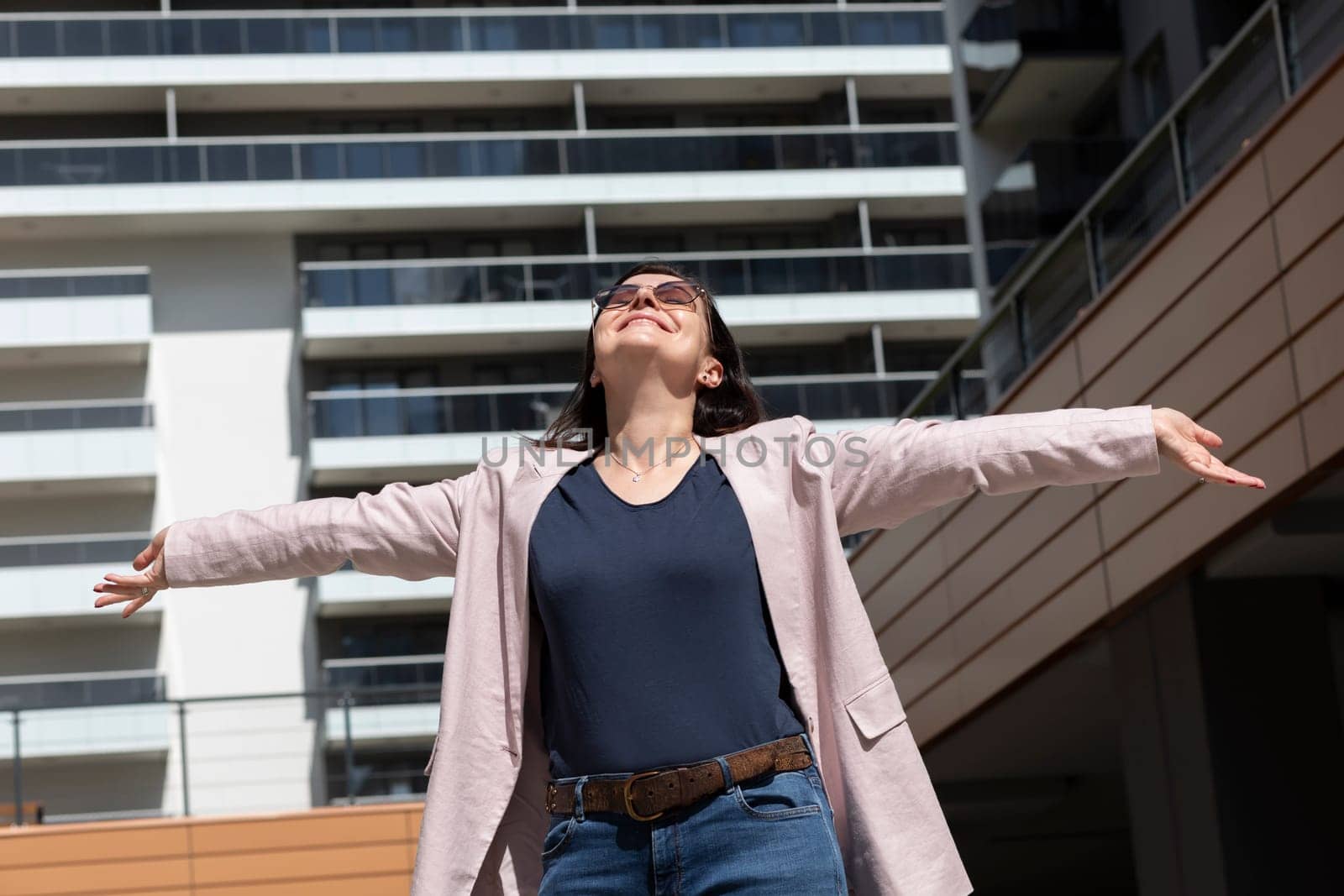 Happy Mature Woman With Open Arms Enjoys Modern Residential Building, Standing In New Apartment Campus, Complex In Sunny Day. Female Brunette Wears Casual Cloth, Horizontal, Real Estate Development.