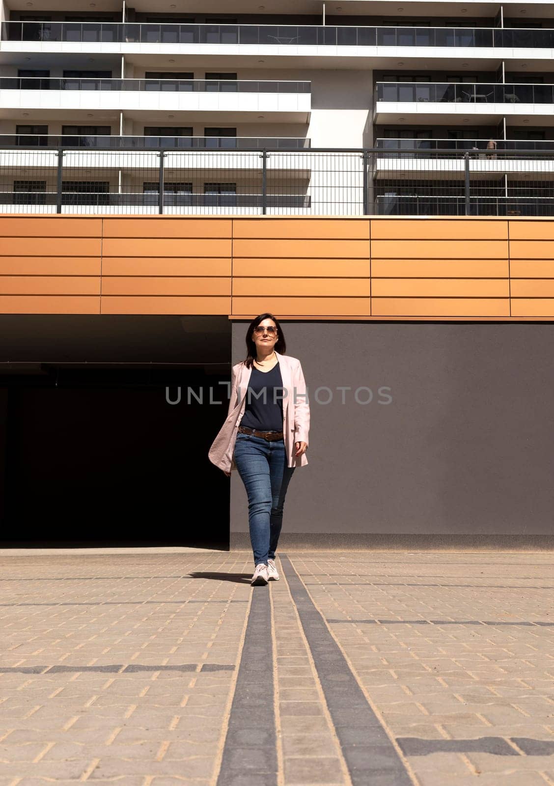 Happy Mature 40 yo Woman Walks In Parking Of New Apartment Campus, Complex In Sunny Day. Female Brunette Wears Casual Cloth. Construction, Modern Residential Building. Vertical Plane.