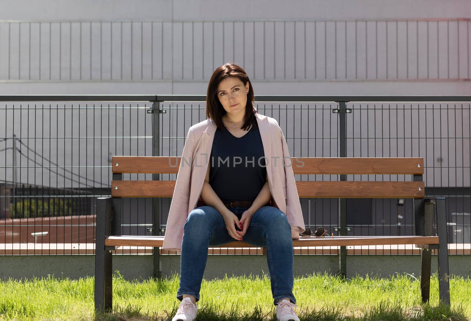 Beautiful Brunette Mature Woman Sitting On A Bench In Residential Campus. Confident Female Is Dressed In Casual Clothes. Rest And Relaxation. Horizontal Plane. High quality photo