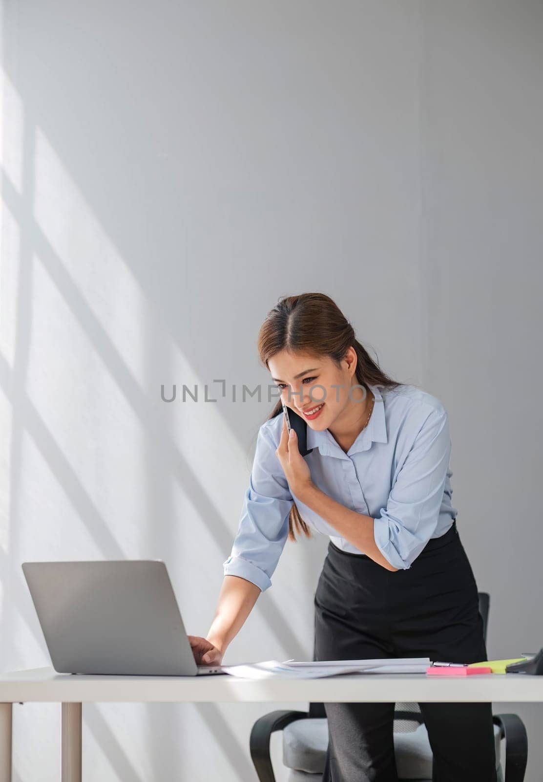 Attractive brunette Asian woman using mobile phone, smile and happy while standing at working desk in modern office