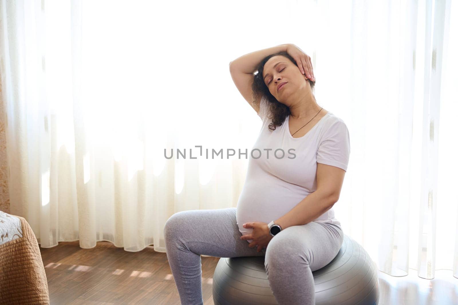 Portrait of multi ethnic pregnant cute woman, expectant gravid mother holding hand on her belly, doing stretching exercises, working out at home, sitting on a fitball. Pregnancy. Maternity lifestyle