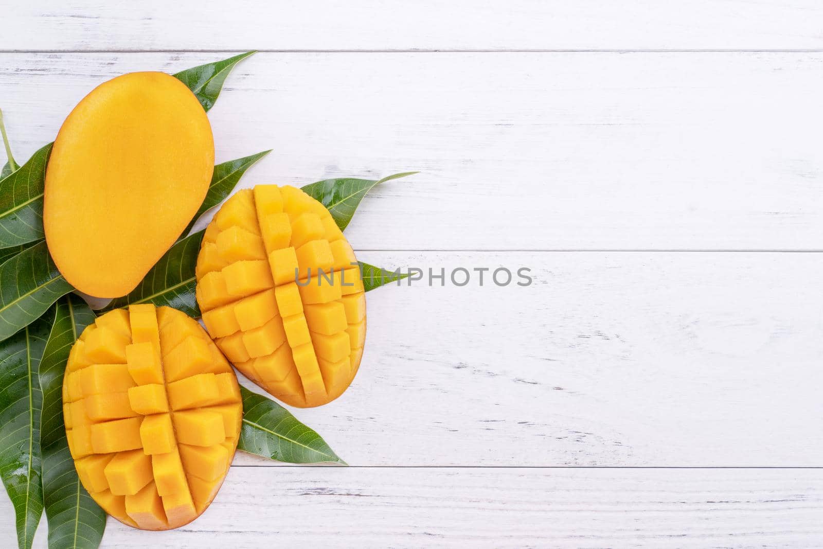Beautiful chopped ripe mango with green leaves on bright white color wooden background, top view, flat lay, copy space. Tropical fruit design concept.