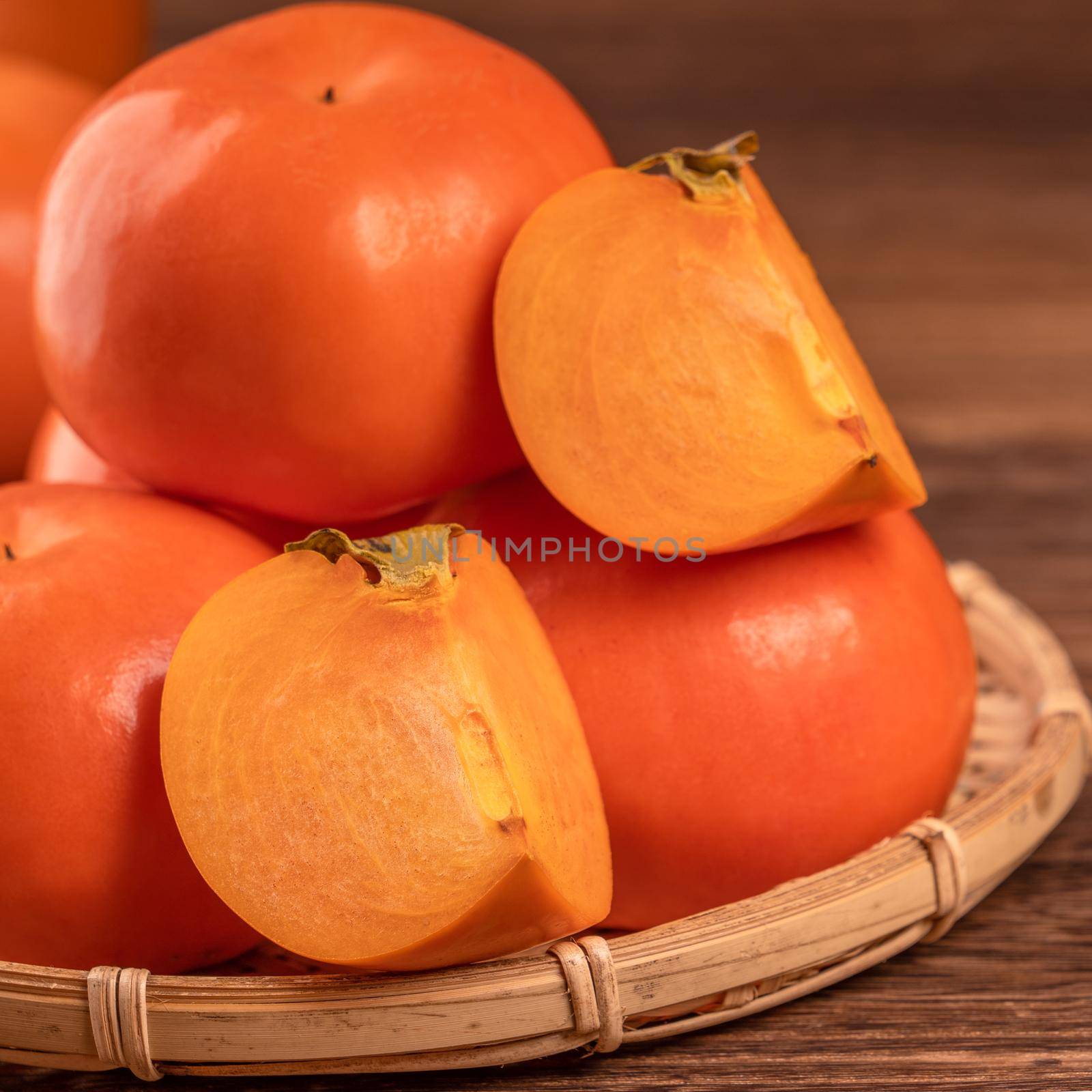 Sliced sweet persimmon kaki in a bamboo sieve basket on dark wooden table with red brick wall background, Chinese lunar new year fruit design concept, close up. by ROMIXIMAGE