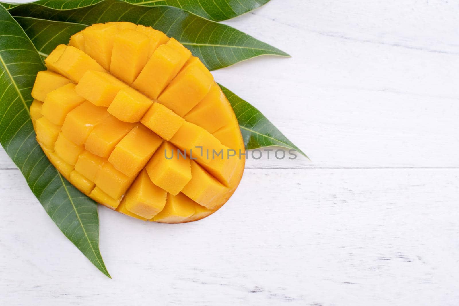 Beautiful chopped ripe mango with green leaves on bright white color wooden background, top view, flat lay, copy space. Tropical fruit design concept.