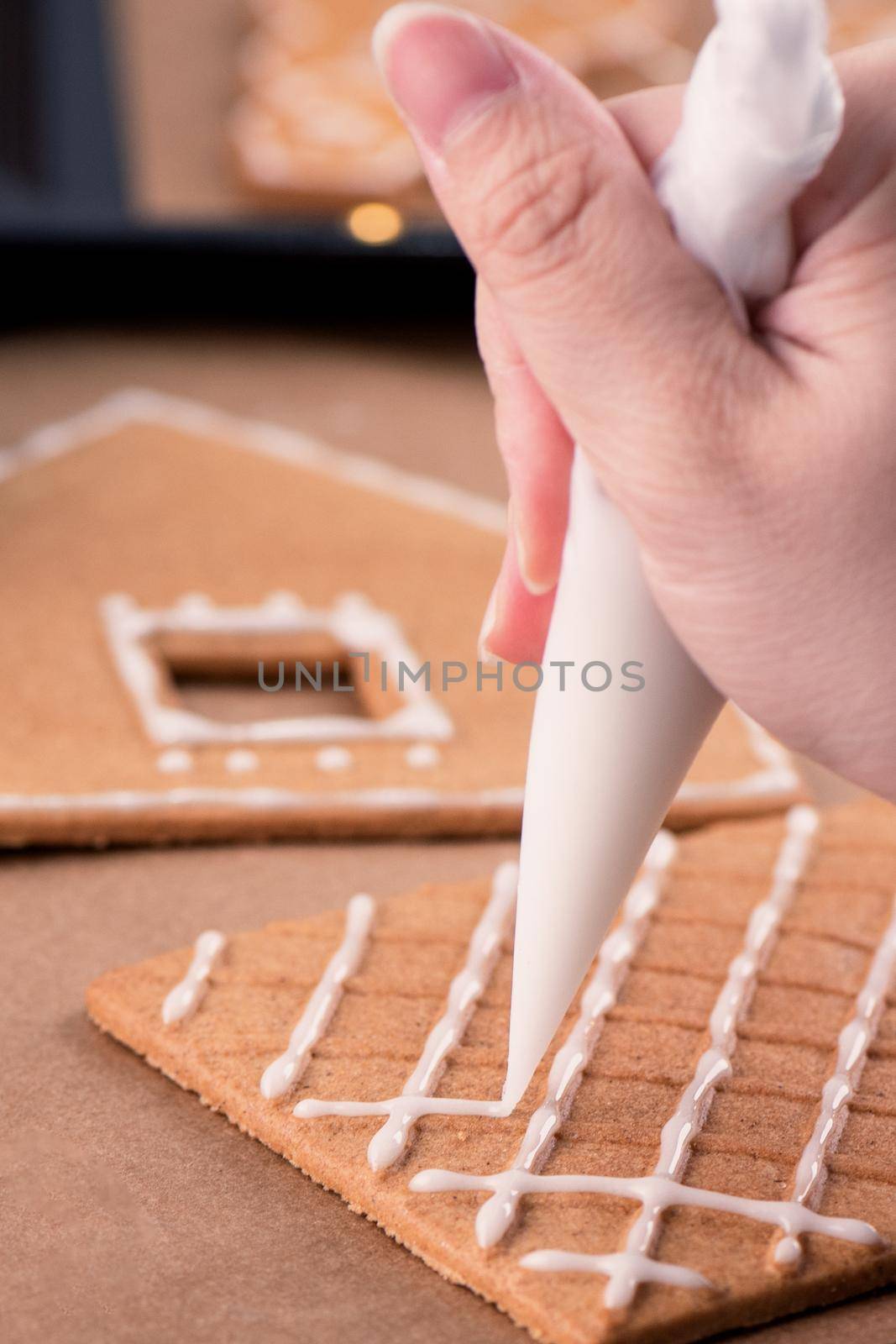 Woman is decorating gingerbread cookies house with white frosting icing cream topping on wooden table background, baking paper in kitchen, close up, macro.