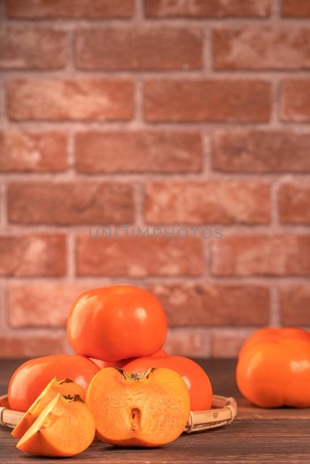 Sliced sweet persimmon kaki in a bamboo sieve basket on dark wooden table with red brick wall background, Chinese lunar new year fruit design concept, close up.