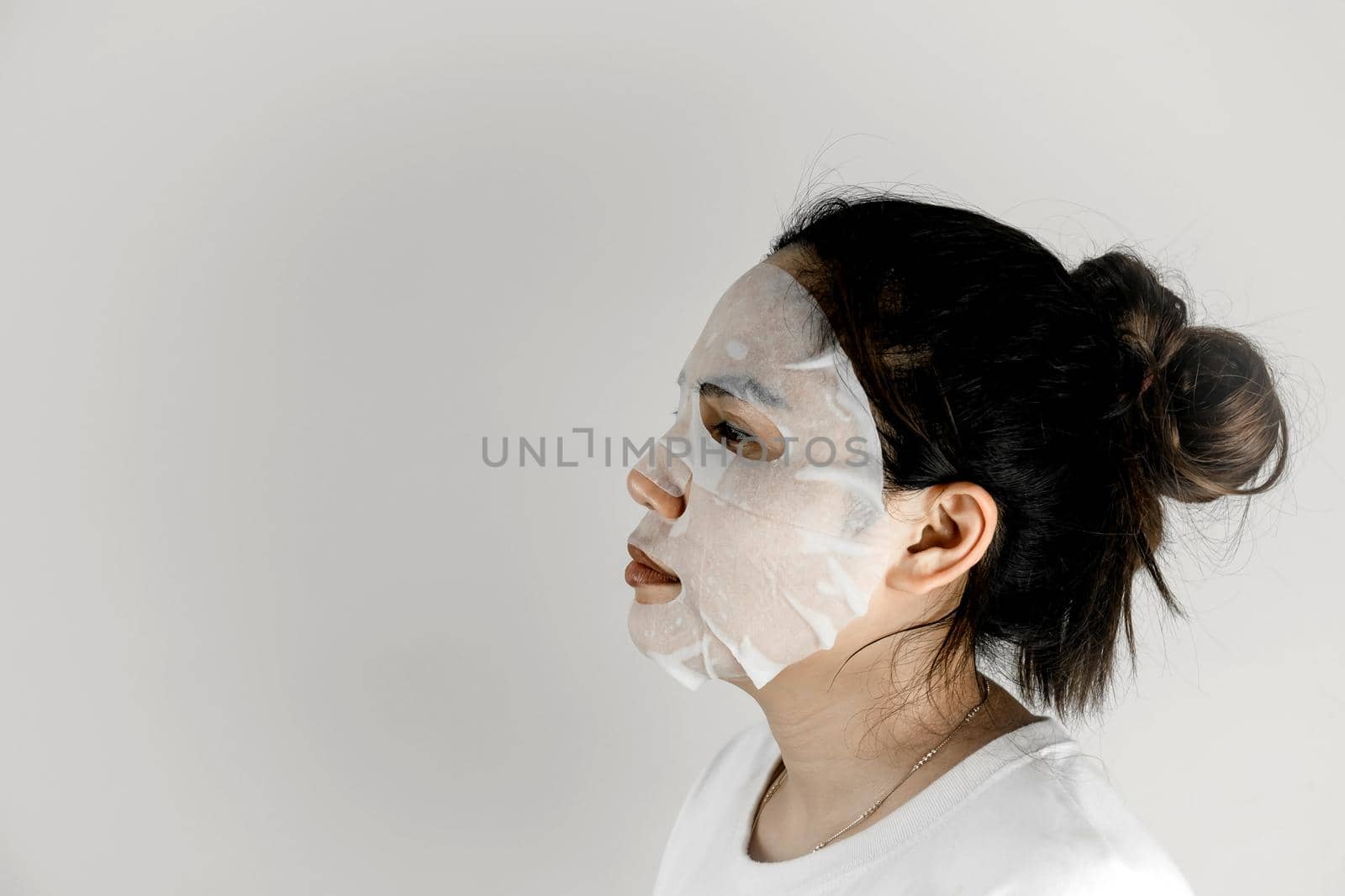 Side view of Asian woman wearing white t-shirt and covering her face with a sheet mask.