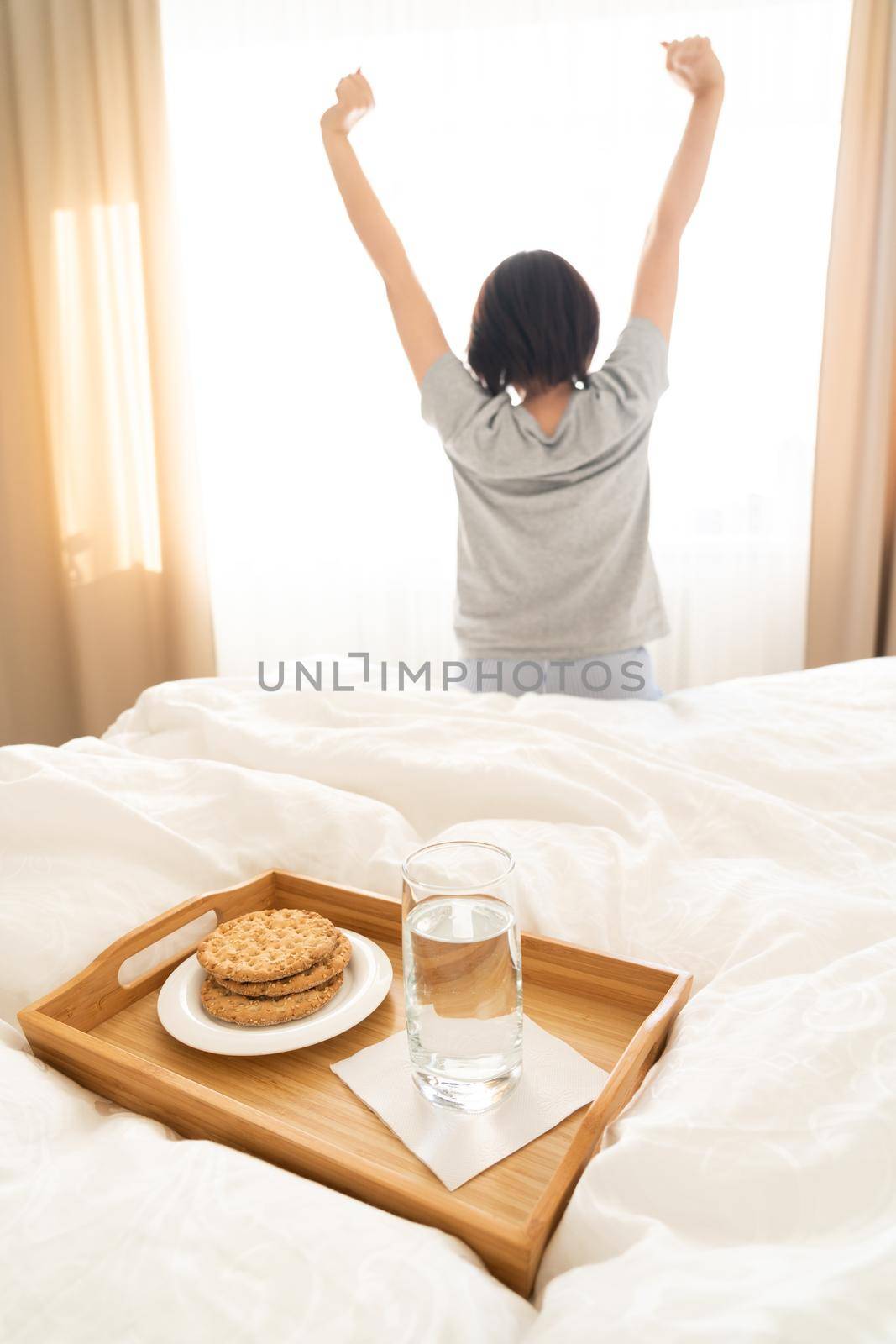 Tray with water and crackers breakfast on a bed with woman stretching on background by Mariakray