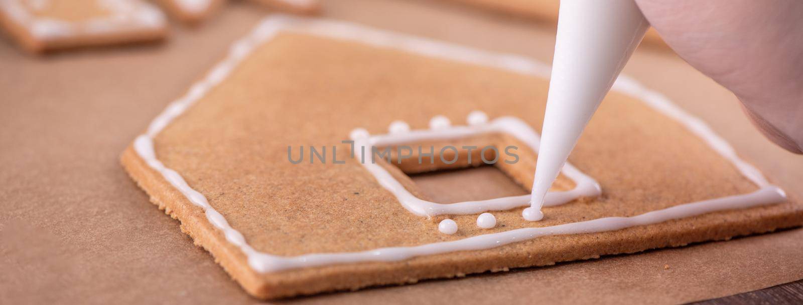 Woman is decorating gingerbread cookies house with white frosting icing cream topping on wooden table background, baking paper in kitchen, close up, macro.