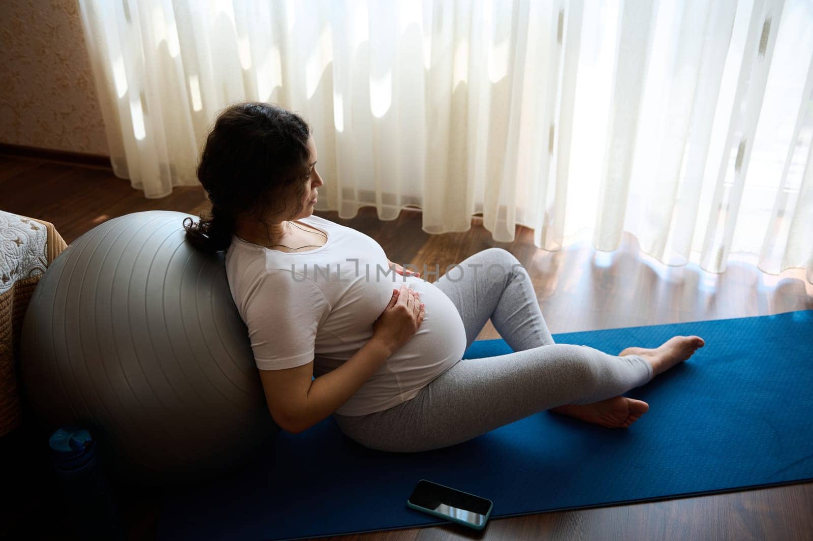 Authentic portrait of delightful pregnant woman, expectant mother holding her hands on her big belly, relaxing leaning on a fit ball after prenatal fitness at home, enjoying tranquil homely atmosphere