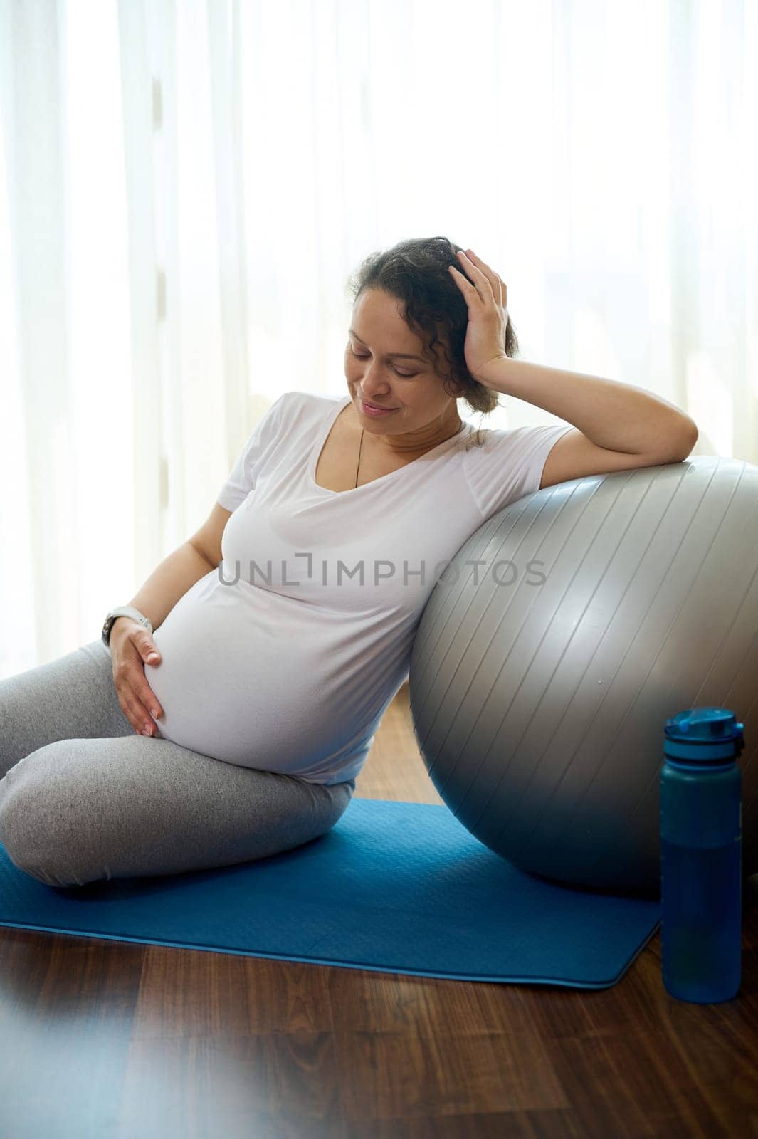 Happy pregnant woman gently stroking her belly, relaxing on a yoga mat after prenatal stretching exercises on fitball, smiling enjoying a healthy active lifestyle in pregnancy time and maternity leave
