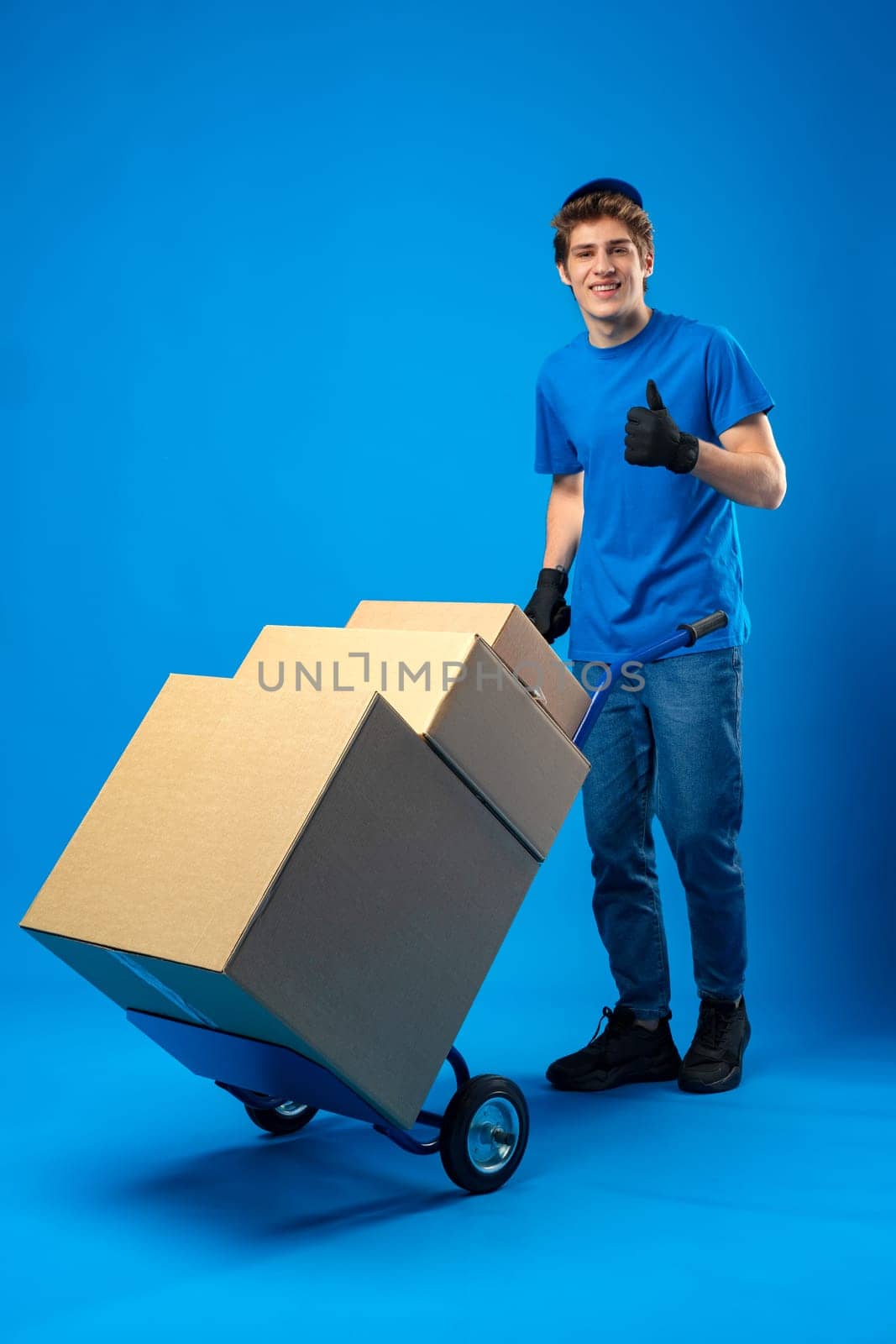 Young happy delivery man carrying boxes on truck against blue background in studio