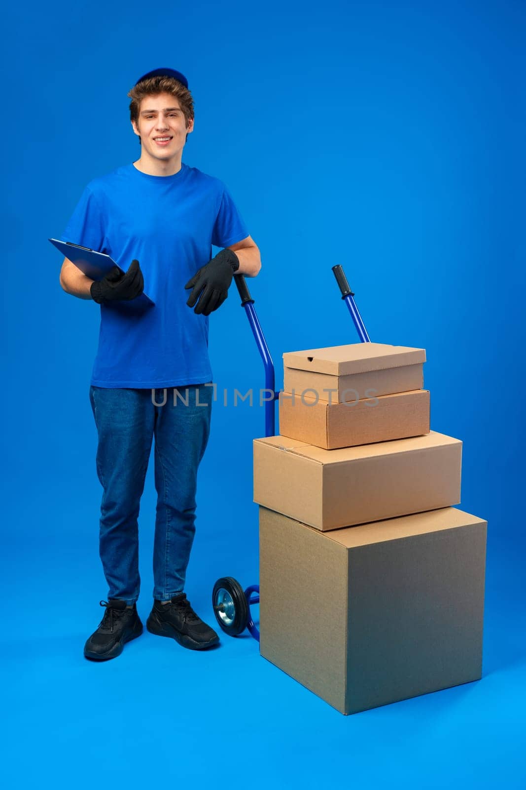 Young happy delivery man carrying boxes on truck against blue background in studio