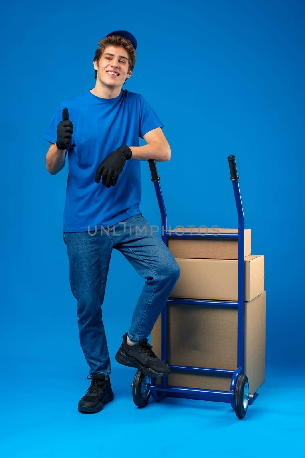 Young happy delivery man carrying boxes on truck against blue background in studio